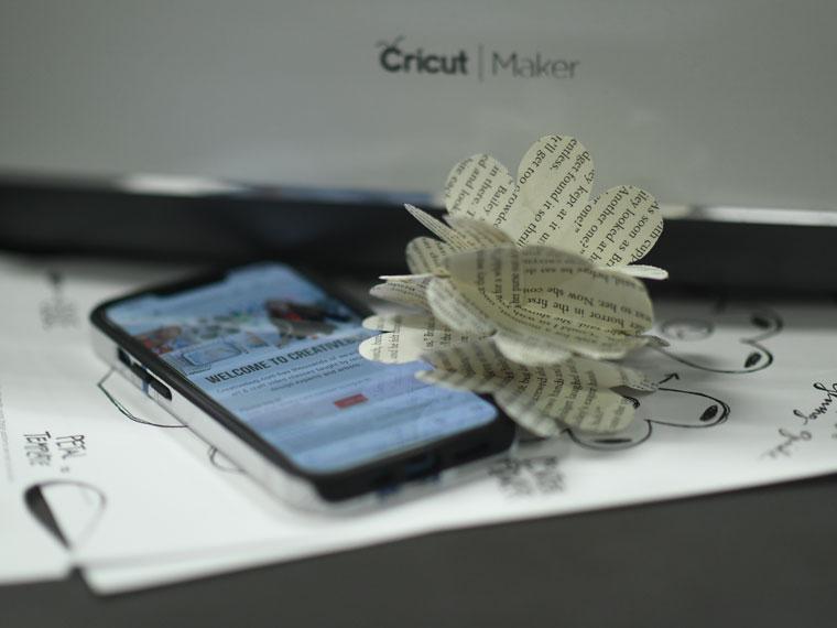 Mobile phone with Creativebug in front of a Cricut machine with a paper flower.