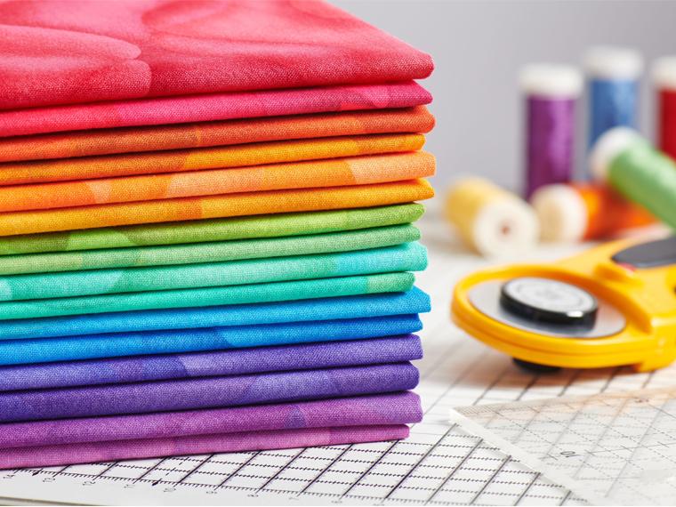 A photograph of a stack of colour fabric sitting on a table. Thread and a rotray cutter sit beside.