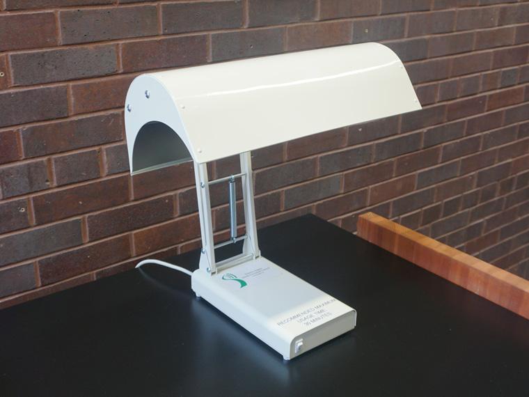 A white light therapy lamp on a black desk, with a brick wall in the background 