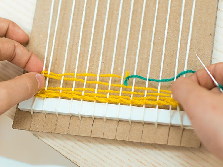 a photograph of hands weaving yellow yarn through a cardboard loom