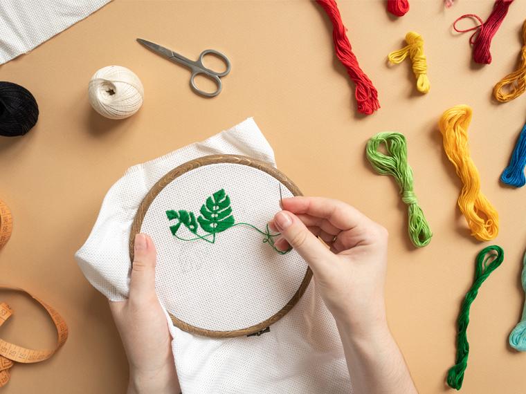 A photograph of a hand stitching a leaf with green embroidery thread on a off white piece of fabric.