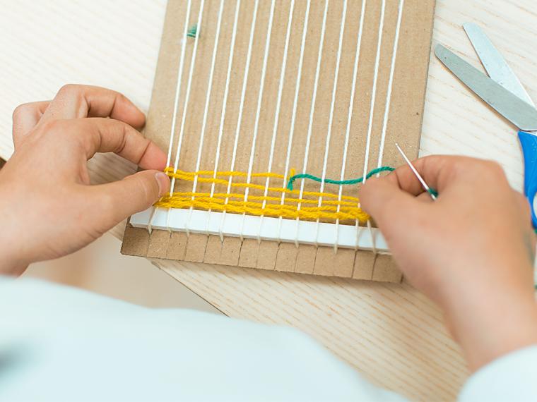 a photograph of hands weaving yellow yarn through a cardboard loom