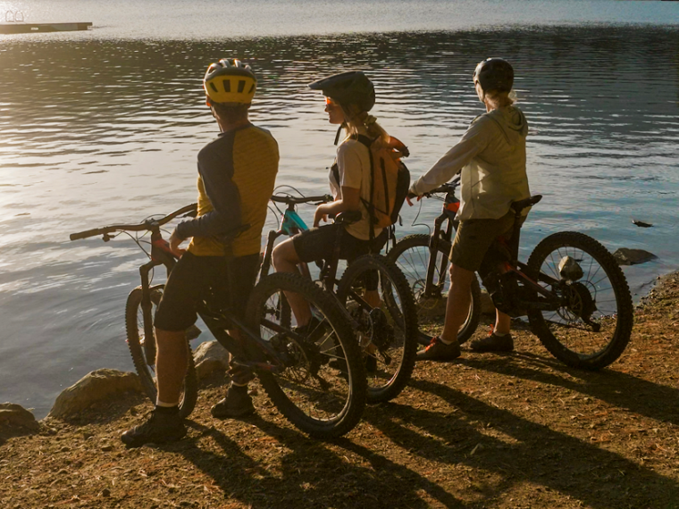 Three people with mountain bikes pause by a lakeshore at sunset.