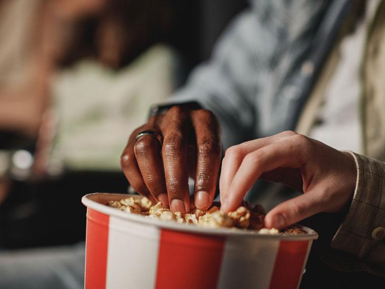 Close up of a couple's hands getting popcorn put of a bucket while watching a movie.