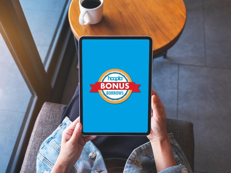 Top view of a person sitting at a table holding an iPad with hoopla Bonus Borrow logo on the screen.