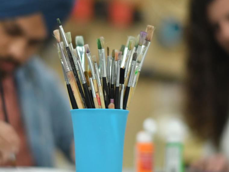 Close up of paint brushes in a cub with two people painting in the background.