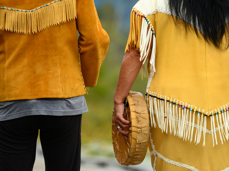 Two First Nations people standing together, wearing traditional clothing and carrying a hide drum