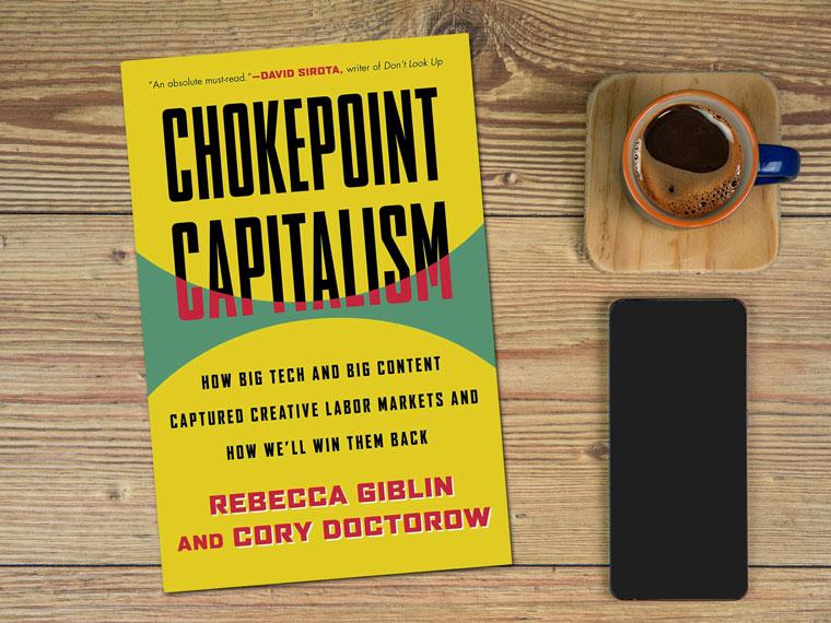 Copy of the book Chokepoint Capitalism on a wooden desk with a cell phone and coffee next to it.