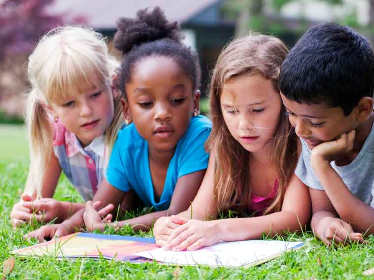 Four children read a book together on the grass in a park.