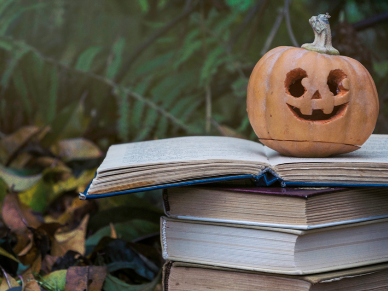 Ripe Halloween carved pumpkin and a stack of old books on the background of fallen autumn yellow leaves in the garden.