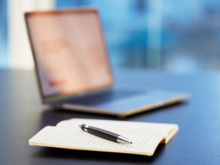 Notebook and pen on a desk with a laptop in the background.