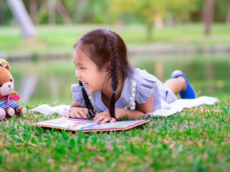 Child lying on grass, laughing, "reading" a book to stuffed animals