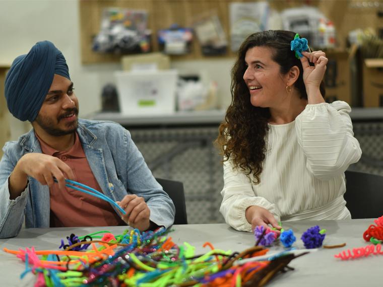 A photograph showing two people sitting at a table making colourful pipecleaner flowers. The person on the left is male-presenting and wears a open denim shirt over a salmon-coloured polo shirt. He wears a navy blue turban and is holding two bright blue pipecleaners. The person on the right is female-presenting. They have long brown curly hair and are wearing a white blouse. She is in the middle of tucking a blue pipecleaner flower behind her ear. 