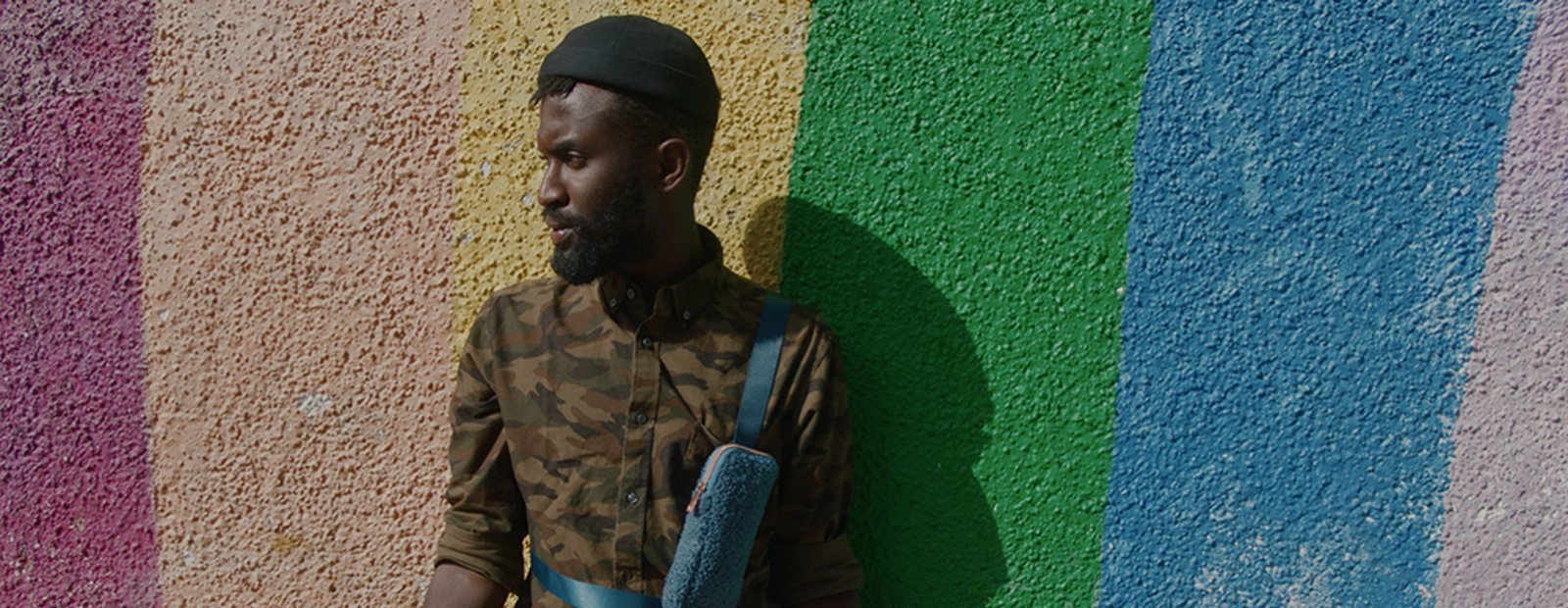 An adult standing outside next to a rainbow-painted wall.