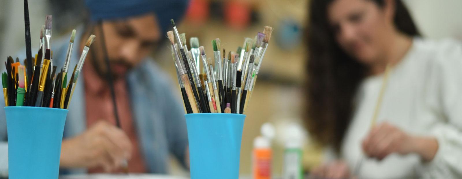 Close up of paint brushes in a cub with two people painting in the background.