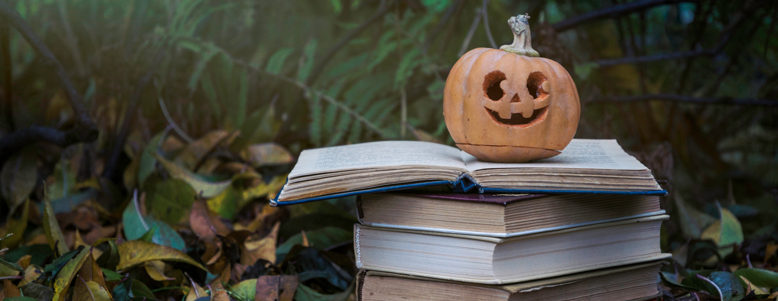 Ripe Halloween carved pumpkin and a stack of old books on the background of fallen autumn yellow leaves in the garden.