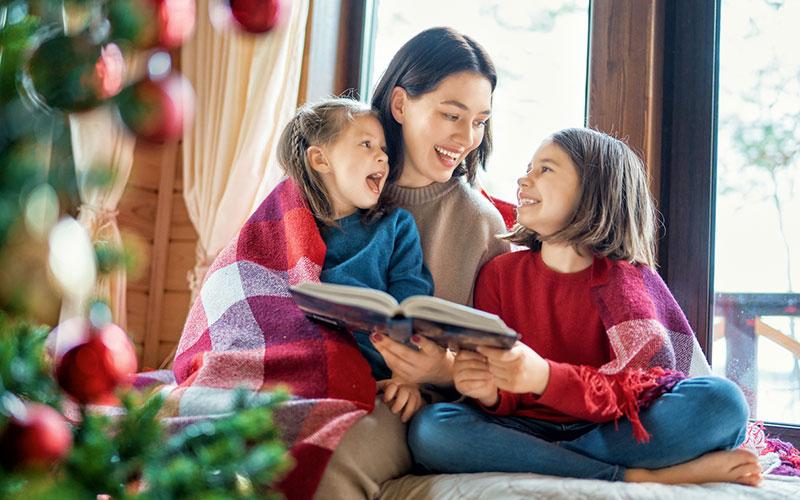 Adult and two kids reading a book next to a Christmas tree.