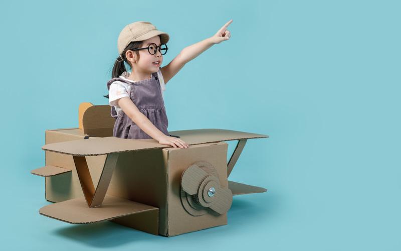 A child stands in a cardboard plane with an arm extended forward.
